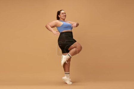 Photo for Young, positive woman with overweight figure training in sportswear against brown studio background. Cardio exercises. Concept of sport, body-positivity, weight loss, body and health care. Ad - Royalty Free Image