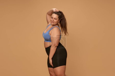 Photo for Portrait of young, positive, overweight woman posing in comfortable sportswear, leggings and top against brown studio background. Concept of sport, body-positivity, weight loss, body and health care - Royalty Free Image