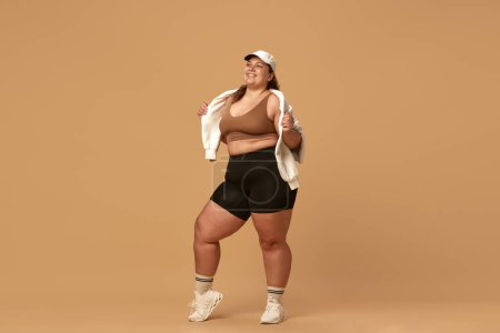 Photo for Young, positive, smiling, overweight woman posing in stylish, trendy sportswear with cap against brown studio background. Concept of sport, body-positivity, weight loss, body and health care - Royalty Free Image
