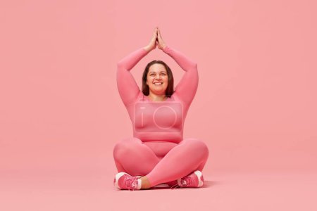 Foto de Yoga y meditación, bienestar. Entrenamiento de mujer joven con sobrepeso en ropa deportiva contra fondo de estudio rosa. Salud mental. Concepto de deporte, positividad corporal, pérdida de peso, cuidado corporal y de la salud - Imagen libre de derechos