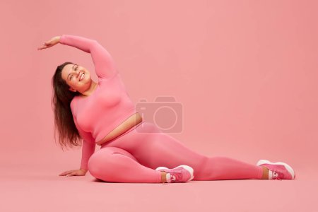 Bien-être et amour-propre. Jeune femme en surpoids s'entraînant en vêtements de sport roses, en faisant des étirements sur fond de studio rose. Concept de sport, positivité corporelle, perte de poids, corps et soins de santé