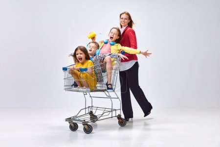 Photo for Portrait of happy young woman, mother going shopping with her little kids, children sitting in shopping trolley against grey studio background. Concept of family, motherhood, childhood, lifestyle - Royalty Free Image