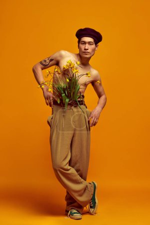 Photo for Full-length portrait of handsome, stylish, young man posing shirtless in beige pants against yellow studio background. Blooming flowers. Concept of mens fashion, style, art, beauty - Royalty Free Image