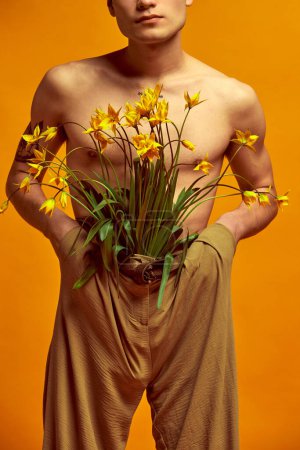 Photo for Spring fashion trends. Cropped image of man posing shirtless in beige classical pants with sticking out flowers against yellow studio background. Concept of mens fashion, style, art, beauty - Royalty Free Image