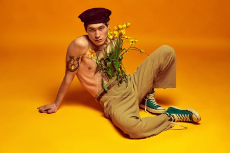 Téléchargez les photos : Ambiance jeunesse. Beau, élégant, jeune coréen posant torse nu avec des fleurs, en béret et pantalon sur fond de studio jaune. Concept de mode masculine, style, art, beauté, tendances - en image libre de droit