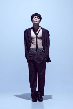 Téléchargez les photos : Portrait de jeune homme élégant posant en veste noire sur corps nu avec cravate et pantalon classique sur fond studio bleu clair. Concept de mode masculine, style, art, beauté, tendances - en image libre de droit