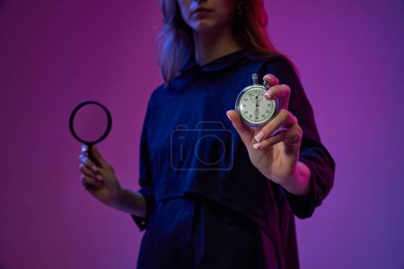 Foto de Gestión del tiempo e investigación. Mujer joven sosteniendo reloj y lupa sobre fondo púrpura degradado en luz de neón. Concepto de negocio, tecnologías modernas, innovaciones - Imagen libre de derechos