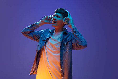 Foto de Retrato de hombre joven relajado en ropa casual, escuchando música en auriculares, divirtiéndose contra el gradiente fondo púrpura en luz de neón. Concepto de emociones humanas, estilo de vida, juventud - Imagen libre de derechos
