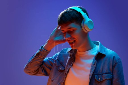 Photo for Portrait of young, smiling, positive man in casual clothes, listening to music in headphones against gradient purple background in neon light. Concept of human emotions, lifestyle, youth - Royalty Free Image