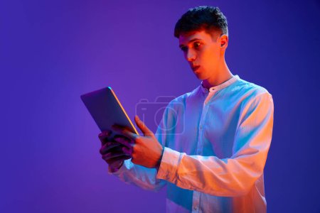 Photo for Portrait of young guy in white shirt emotionally looking in tablet against gradient purple background in neon light. Betting, success, winning. Concept of human emotions, lifestyle, youth - Royalty Free Image