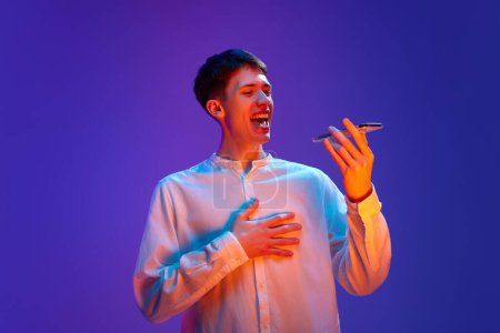 Photo for Portrait of young man in white shirt emotionally recording voice message on phone, talking against gradient purple background in neon light. Online communication. Concept of human emotions, lifestyle - Royalty Free Image