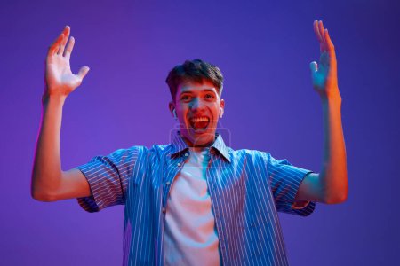 Photo for Portrait of young guy in casual clothes, with headphones, spreading hands in happiness and excitement against gradient purple background in neon light. Concept of human emotions, lifestyle, youth - Royalty Free Image
