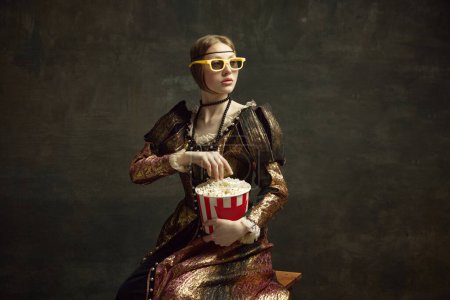 Téléchargez les photos : Portrait de princesse médiévale en robe vintage et lunettes de film 3D, mangeant du pop-corn sur fond vert foncé. Concept d'histoire, remake de l'art de la Renaissance, comparaison des époques, loisirs modernes - en image libre de droit