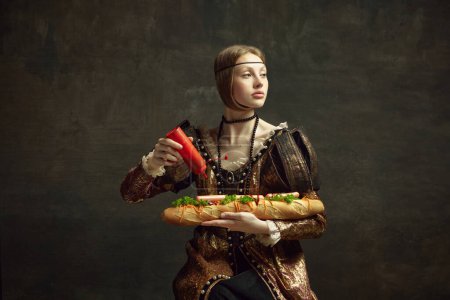 Foto de Retrato de niña, reina, princesa en traje vintage poniendo ketchup en baguette sándwich gigante sobre fondo verde oscuro. Concepto de historia, arte renacentista, comparación de épocas, salud y alimentación - Imagen libre de derechos