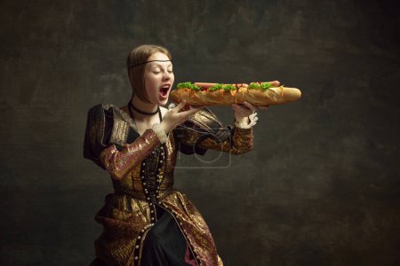 Foto de Retrato de niña, princesa en traje vintage comiendo sándwich de baguette gigante sobre fondo verde oscuro. Concepto de historia, arte renacentista, comparación de épocas, salud y alimentación, dieta - Imagen libre de derechos