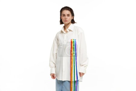 Foto de Retrato de una joven mujer seria en camisa blanca con línea de arco iris que simboliza el apoyo lgbt. Modelo posando sobre fondo blanco. Concepto de comunidad lgbt, apoyo, amor, derechos humanos, mes del orgullo - Imagen libre de derechos