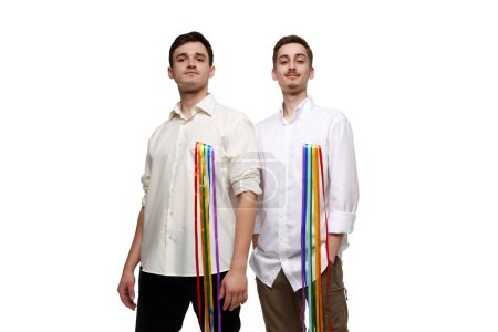 Foto de Retrato de dos jóvenes, orgullosos y confiados, posando en camisa blanca con arco iris, bandera lgbt sobre fondo blanco. Concepto de comunidad lgbt, apoyo, amor, derechos humanos, mes del orgullo - Imagen libre de derechos
