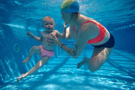 Téléchargez les photos : Petite, drôle, mignonne petite fille, enfant nageant sous l'eau dans la piscine avec mère. Activité familiale. Apprendre à nager. Concept de sport, mode de vie sain et actif, enfance, plaisir et entraînement - en image libre de droit