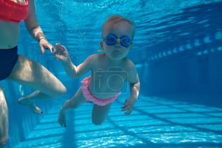Foto de Niña linda, divertida, pequeña, niño pequeño con gafas nadando bajo el agua en la piscina con instructor. Actividad deportiva acuática. Concepto de deporte, estilo de vida saludable y activo, infancia, diversión, entrenamiento - Imagen libre de derechos