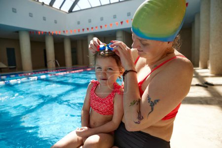 Photo for Smiling happy mother putting goggles on her little beautiful daughter, kid. Swimming in swimming pool indoors. Concept of sport, healthy and active lifestyle, childhood, fun and training - Royalty Free Image