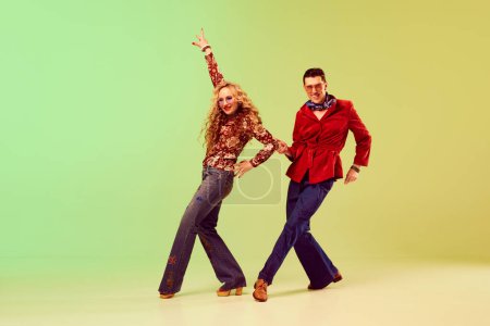 Artistic people. Young couple, man and woman in stylish retro clothes dancing against gradient green yellow background. Concept of retro style, disco dance, fashion, art, hobby, music, 70s