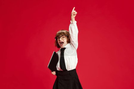 Photo for Portrait of happy, excited little school girl, child in uniform with tablet raising hand against red studio background. Active kid. Concept of childhood, education, fashion, kids emotions - Royalty Free Image
