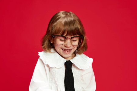 Téléchargez les photos : Portrait de heureuse petite écolière en chemisier blanc avec cravate et gros verres, riant, souriant sur fond de studio rouge. Concept d'enfance, éducation, mode, émotions enfantines - en image libre de droit