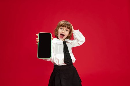 Portrait of happy, smiling, laughing little girl in white blouse and black shirt, showing mobile phone screen against red studio background. Concept of childhood, education, fashion, kid emotions
