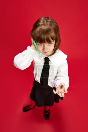 Photo for Full-length top view portrait of little girl in black shirt and white blouse talking on mobile phone with anger against red studio background. Concept of childhood, education, fashion, kid emotions - Royalty Free Image