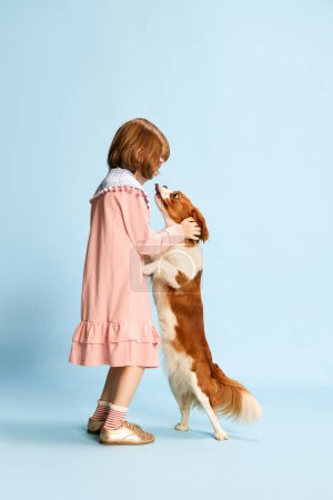 Téléchargez les photos : Portrait complet de petite fille, enfant en robe rose jouant avec un beau chien sur fond de studio bleu. Mon ami. Concept d'enfance, émotions, plaisir, mode, style de vie, animaux domestiques - en image libre de droit