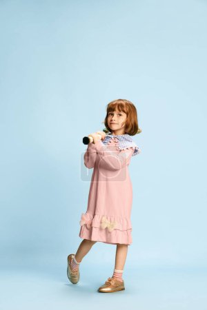 Foto de Retrato de larga duración de una niña sonriente, feliz y pequeña en vestido rosa posando con raqueta de tenis sobre fondo azul del estudio. Concepto de infancia, emociones, diversión, moda, estilo de vida activo, deporte - Imagen libre de derechos