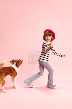 Foto de Retrato de larga duración de la niña feliz, niño en boina roja y suéter a rayas jugando con el perro contra el fondo del estudio rosa. Concepto de infancia, emociones, diversión, moda, estilo de vida - Imagen libre de derechos