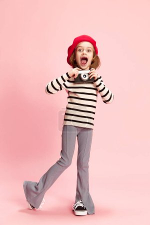 Foto de Retrato de niña feliz, emocionada, cild en suéter a rayas y boina roja posando con camra foto vintage agaisnt fondo de estudio rosa. Concepto de infancia, emociones, diversión, moda, estilo de vida - Imagen libre de derechos