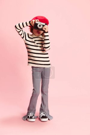 Foto de Retrato de niña, cild en suéter a rayas y boina roja posando con camra foto vintage agaisnt fondo de estudio rosa. Turista. Concepto de infancia, emociones, diversión, moda, estilo de vida - Imagen libre de derechos