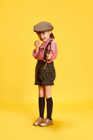 Téléchargez les photos : Portrait de petite fille, enfant en vêtements rétro, chemise et short, tenant fronde sur fond de studio jaune. Concept d'enfance, émotions, plaisir, mode, mode de vie. Jeu de plein air - en image libre de droit