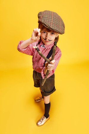 Photo for Portrait of little girl, child in retro style clothes, cap, shirt and shorts, playing with slingshot against yellow studio background. Concept of childhood, emotions, fun, fashion, lifestyle - Royalty Free Image