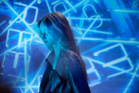 Foto de Una mirada reflexiva. Retrato de joven hermosa chica en chaqueta de cuero posando sobre fondo azul con elementos de neón abstractos. Concepto de arte, estilo moderno, cyberpunk, futurismo y creatividad - Imagen libre de derechos