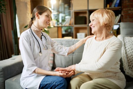 Smiling young woman, doctor supporting her elderly female patient, holding her hand and cheering up. Senior lady at home. Concept of medical care, medicine, illness, health care, profession