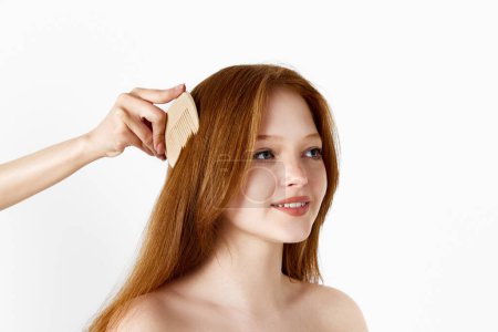 Téléchargez les photos : Femme brossant les cheveux de belle fille rousse tendre sur fond de studio blanc. Soins capillaires cosmétiques. Concept de beauté naturelle, soins capillaires, traitement, cosmétologie, soins de la peau - en image libre de droit