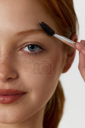 Photo for Close-up portrait of beautiful young redhead female model with freckles, brushing brows against white studio background. Concept of natural beauty, cosmetology, cosmetics, skin care - Royalty Free Image
