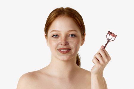 Photo for Portrait of attractive young, redhead girl with naked shoulders, holding eyelash curler against white studio background. Freckled face. Concept of natural beauty, cosmetology, cosmetics, skin care - Royalty Free Image