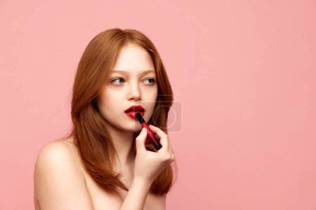 Téléchargez les photos : Portrait de jeune, belle fille rousse à la peau bien entretenue, appliquant du rouge à lèvres rouge sur fond de studio rose. Concept de beauté naturelle, maquillage, cosmétologie, cosmétique, soins de la peau - en image libre de droit
