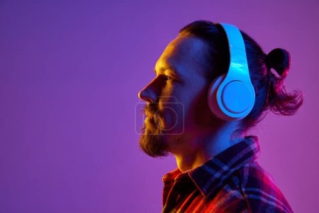 Photo for Side view portrait of young man listening to music in headphones against purple studio background in neon light. Concept of human emotions, facial expression, lifestyle. Copy space for ad - Royalty Free Image