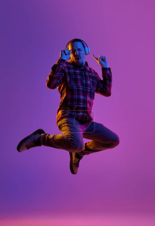 Photo for Portrait of emotional man in casual clothes, listening to music in headphones and jumping against purple studio background in neon light. Concept of human emotions, facial expression, lifestyle - Royalty Free Image