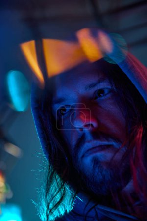 Photo for Portrait of serious man in hoodie attentively looking away against blue studio background in neon light. Night life. Concept of human emotions, facial expression, lifestyle - Royalty Free Image