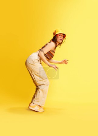 Photo for Full-length portrait of funny young girl in panama, pants and top slinking up with funny face against yellow studio background. Concept of human emotions, youth culture, fashion, lifestyle, meme look - Royalty Free Image