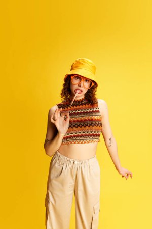Portrait of lovely young girl with curly hair, posing in panama, glasses and knitted top, eating bubble gum over yellow studio background. Concept of human emotions, youth culture, fashion, lifestyle
