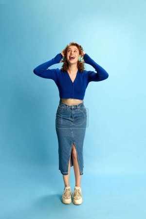 Foto de Retrato de larga duración de una joven feliz y sonriente con top azul y falda de jeans escuchando música en auriculares con fondo de estudio azul. Concepto de emociones humanas, cultura juvenil, moda, música - Imagen libre de derechos