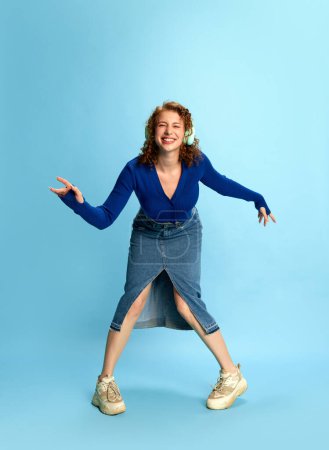 Foto de Retrato de una joven feliz con top azul y falda de jeans escuchando música en auriculares y bailando emocionalmente sobre fondo de estudio azul. Concepto de emociones humanas, cultura juvenil, moda - Imagen libre de derechos