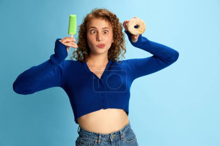 Photo for Portrait of funny looking young curly girl in blue top holding ice cream and donut against blue studio background. Concept of human emotions, youth culture, fashion, lifestyle. Meme face - Royalty Free Image